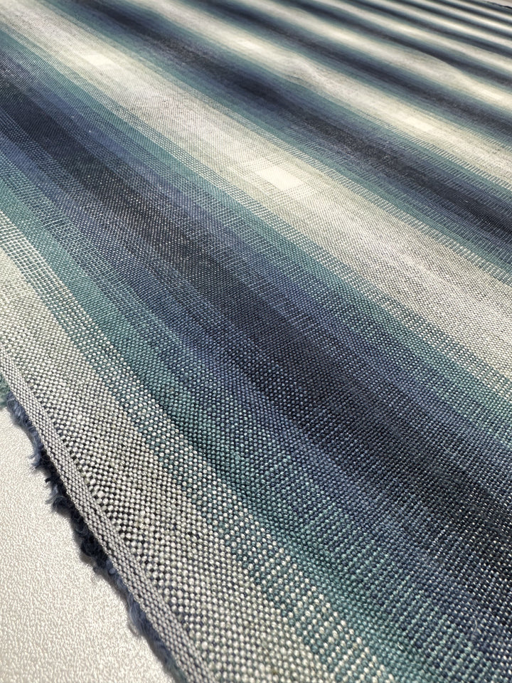 Close-up of Pure Linen - Peacock by Super Cheap Fabrics, showcasing shades of blue, gray, and green. The heavy weight linen features a textured weave with horizontal stripes creating a gradient effect, beautifully enhanced by light to reveal intricate details. Width: 148cm.