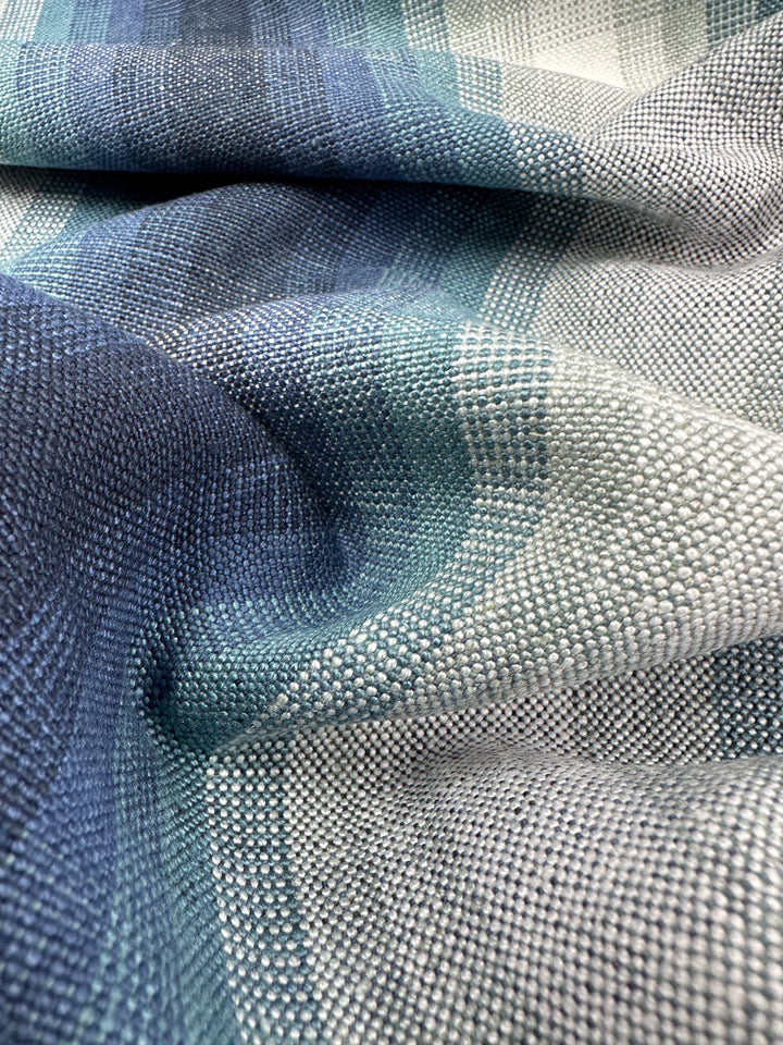 A close-up of Super Cheap Fabrics' Pure Linen - Peacock - 148cm shows a textured upholstery fabric with blue and gray gradient tones. Its intricate weave pattern gives it a soft, flowing look, while light accents shimmer across the surface.