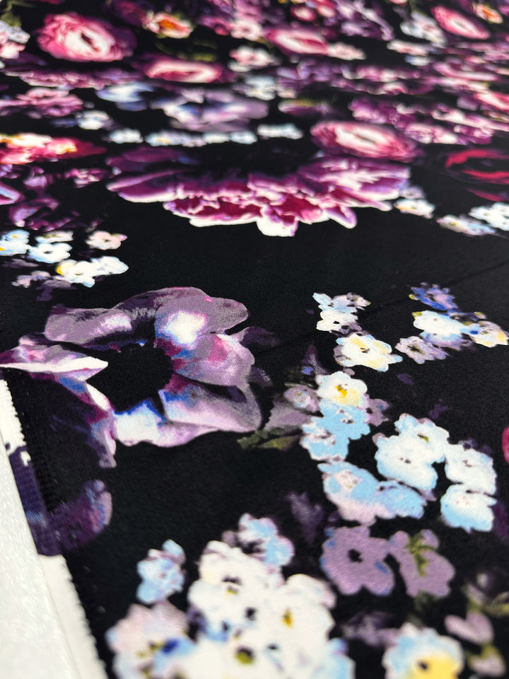 The Cotton Sateen - Arrangement - 125cm from Super Cheap Fabrics features a vivid floral design with large pink and purple flowers, complemented by smaller white and blue blossoms on a black background, ideal for dresses with intricate details and vibrant, striking contrast.