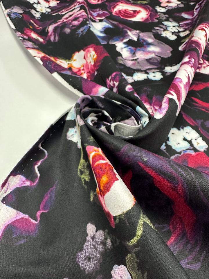 A close-up of Cotton Sateen - Arrangement - 125cm from Super Cheap Fabrics, featuring a black base with vibrant pink, red, and white floral patterns. The partially folded fabric highlights its smooth texture and colorful design, ideal for crafting elegant dresses.