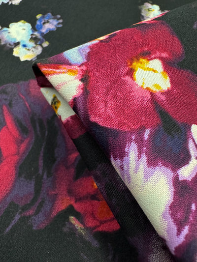The Cotton Sateen - Arrangement by Super Cheap Fabrics features a close-up view of its textured material, displaying vibrant floral patterns with large red, pink, and purple flowers on a dark background. A folded corner highlights the texture, making it ideal for elegant dresses.