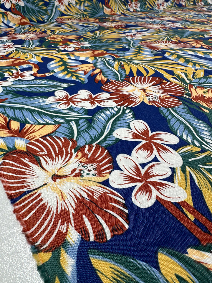 A close-up of the Super Cheap Fabrics' Pure Printed Linen - Lost - 135cm displays a tropical design with large white and red hibiscus flowers, smaller white blooms, and lush green leaves on a rich blue background. Its light weight gives an airy feel to the vibrant pattern.