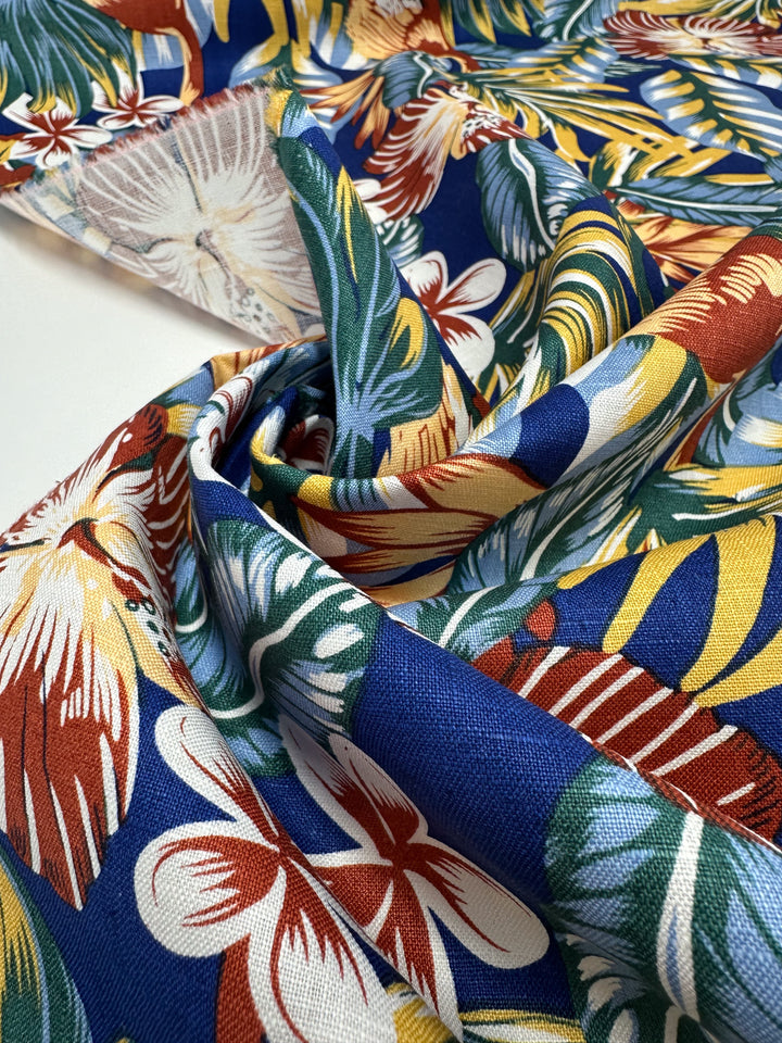 The Pure Printed Linen - Lost by Super Cheap Fabrics is a lightweight, multi-coloured fabric featuring a vibrant tropical print with blue, yellow, green, white, and red hues. Its design includes large leaves and flowers on a slightly wrinkled texture for added depth, measuring 135cm wide.