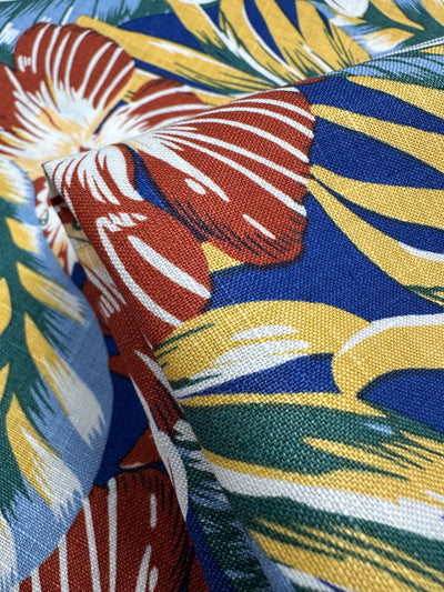 Close-up of Pure Printed Linen - Lost - 135cm by Super Cheap Fabrics, featuring large red, white, and yellow flowers with green leaves on a blue background. The lightweight, vibrant tropical fabric is slightly folded to show its texture and colorful pattern.