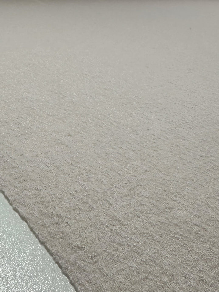 A close-up of the elegant "Boiled Wool - Coconut Milk - 140cm" from Super Cheap Fabrics, showcasing a subtle wavy texture. The muted, light color features a metallic corner in the bottom left reflecting light.