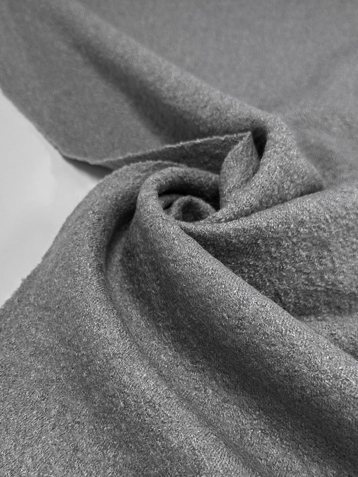 A close-up of the Boiled Wool - Gull - 142cm by Super Cheap Fabrics shows a soft gray wool-viscose blend with a smooth, slightly fuzzy texture. The fabric is draped in gentle folds and curves, creating an elegant spiral design, similar to a neatly arranged piece of cloth.