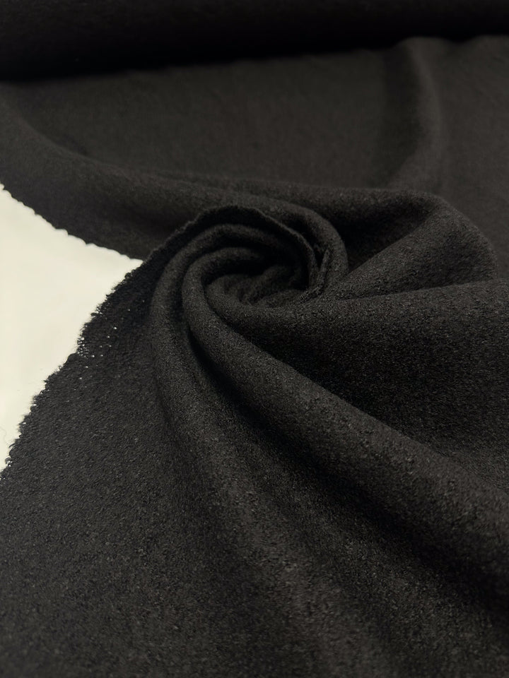 A close-up of Super Cheap Fabrics' Boiled Wool in black, 145cm wide, showcases its soft texture and heavy weight. The fabric is spirally arranged with a slight sheen and visible weave, prominently displayed against a neutral background.