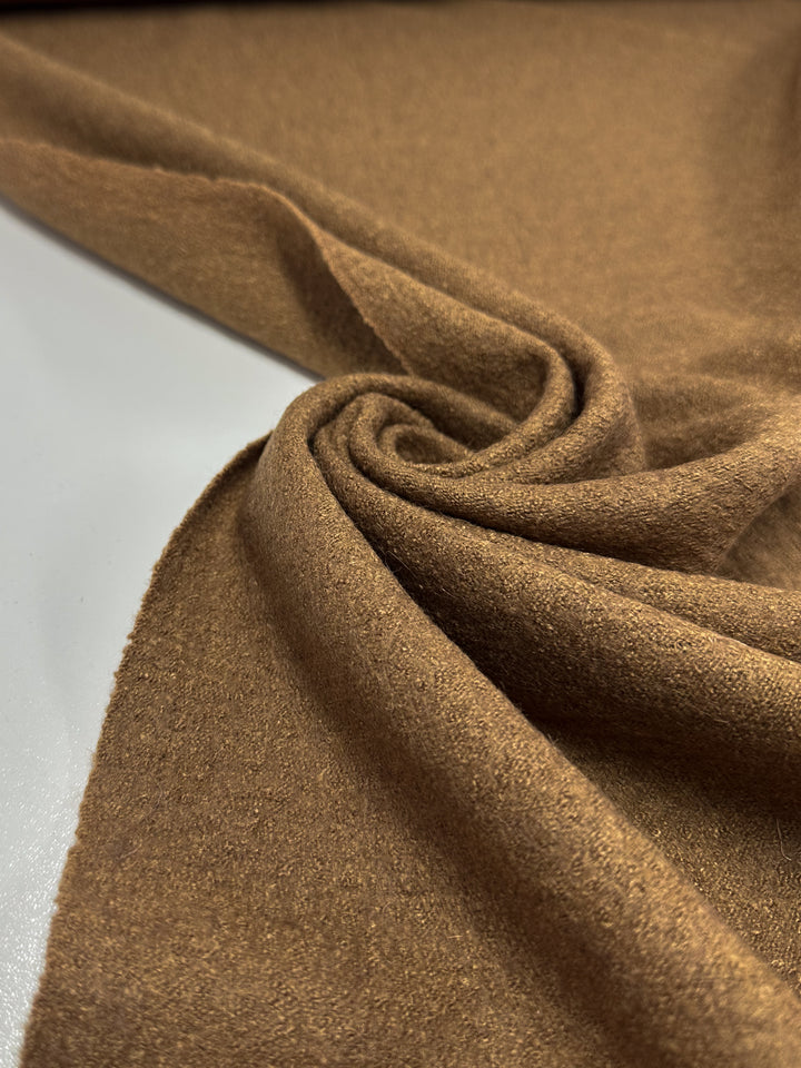 A close-up showcases the texture and folds of the Boiled Wool - Tobacco fabric by Super Cheap Fabrics, with its soft brown wool-viscose blend draped into a gentle swirl on a flat surface. Width: 145cm.