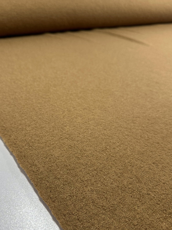 A roll of Super Cheap Fabrics' Boiled Wool in Oak (147cm) elegantly unfurls on a flat surface. Its soft, smooth texture reflects light beautifully, creating subtle highlights and shadows as it extends invitingly into the background.