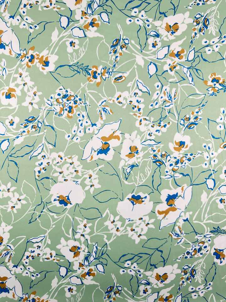 The Printed Stretch Satin - Green Sakura from Super Cheap Fabrics showcases white and orange flowers with green leaves on a light green backdrop, enhanced by blue outlines. This lightweight fabric offers a vibrant, botanical feel reminiscent of printed satins. Width: 150cm.