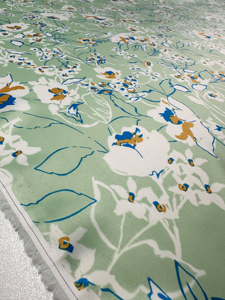 The Printed Stretch Satin - Green Sakura by Super Cheap Fabrics offers a close-up of lightweight fabric in light green, showcasing an intricate floral pattern in white, blue, and orange with various leaves and flowers for a vibrant look. Width: 150cm.