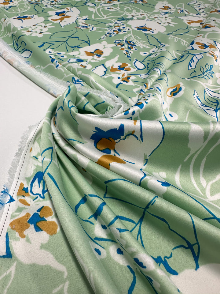 A close-up of elegant pieces in luxurious Printed Stretch Satin - Green Sakura by Super Cheap Fabrics, showcasing vibrant floral patterns in white, yellow, and blue. The fabric's gentle drape enhances its texture and design, with visible selvage on the left.