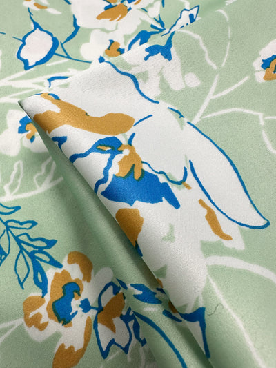 Close-up of Super Cheap Fabrics' Printed Stretch Satin - Green Sakura (150cm), showcasing an elegant floral pattern with white, tan, and blue flowers on green stems and leaves set against a light green background. Slight folds highlight the satin's texture and drape.