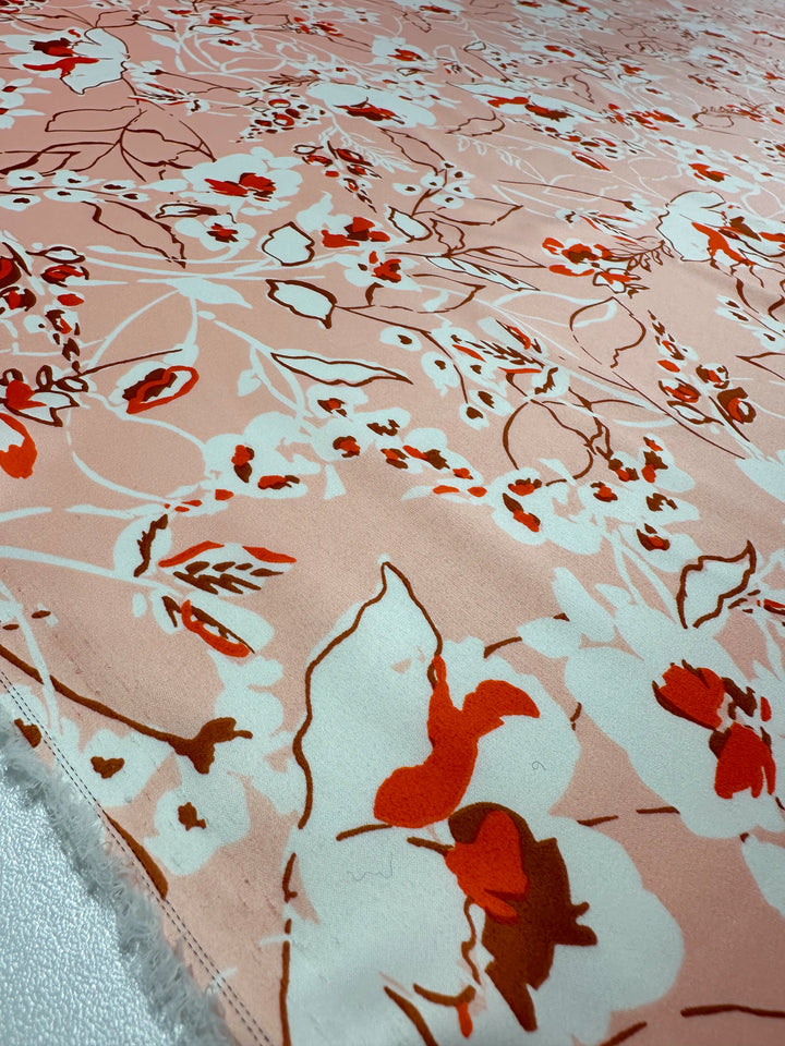 The Printed Stretch Satin - Pink Sakura by Super Cheap Fabrics features a lightweight, peach-based abstract floral design with red, white, and dark brown flowers and leaves arranged randomly across the 150cm fabric.