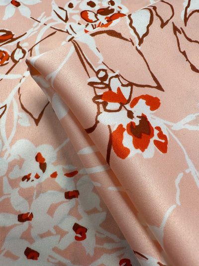 The Printed Stretch Satin - Pink Sakura from Super Cheap Fabrics features a close-up of lightweight, folded fabric on a soft pink background with red and brown floral patterns. The abstract flower shapes and flowing lines give it an elegant, satin-like appearance. Measures 150cm wide.