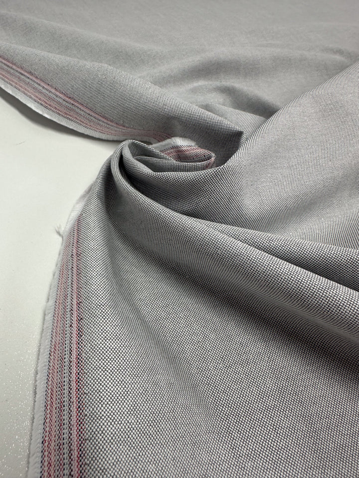A close-up of Super Cheap Fabrics' Linen Blend in Grey White, 150cm, reveals a textured surface with a subtle herringbone pattern and red and white selvedge. The lightweight cotton blend is artistically folded to create elegant curves and shadows.