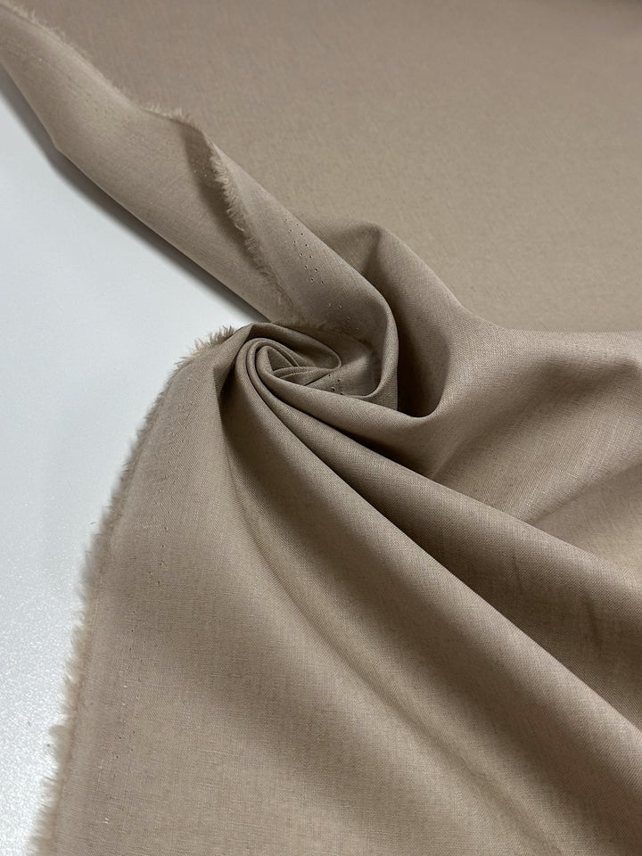 A close-up of the Super Cheap Fabrics Linen Blend - Oxford Tan - 135cm shows light brown linen swirled on a white surface. This lightweight fabric has a soft texture, frayed edges, and is highlighted by lighting that reveals its smooth, slightly matte finish.