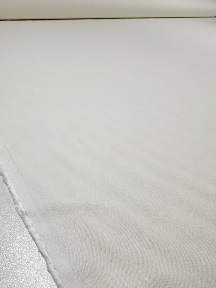 A close-up of Super Cheap Fabrics' "Pure Linen - Ivory - 145cm" showcases its smooth, breathable, soft texture laid flat. The raw edge is slightly frayed, while the shiny surface below accentuates its lightweight appeal.