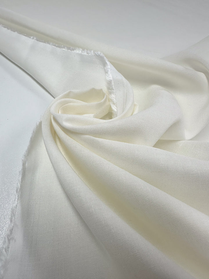A close-up of Super Cheap Fabrics' Pure Linen in Ivory shows its smooth texture and soft folds draped on a light surface. The breathable, fluid qualities are visible, with slightly frayed edges adding to its lightweight charm. Width: 145cm.