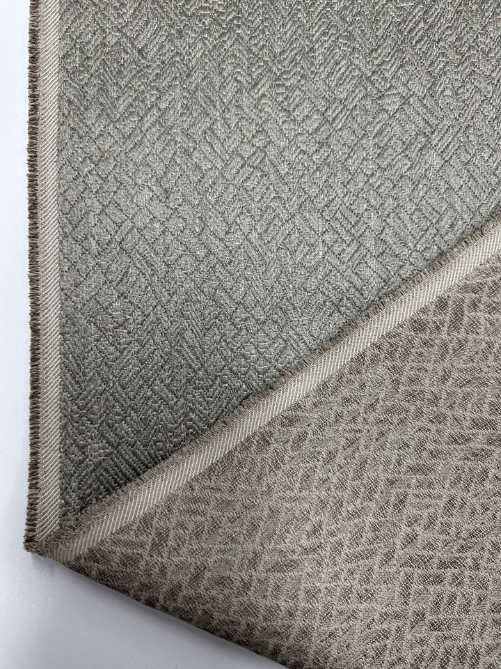 The Herringbone Textured Upholstery by Super Cheap Fabrics in Urbane Silver (width: 143cm) showcases a diagonal cut dividing sections. The top has a darker chevron resembling herringbone, while the lighter bottom features a geometric texture. Neatly trimmed edges add sophistication.