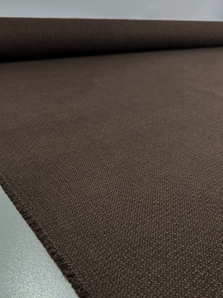 The Basket Weave Upholstery - Tuco by Super Cheap Fabrics is a 147cm wide dark brown fabric with a subtle texture partially unrolled on a flat surface, casting a slight shadow. This heavy-weight material is ideal for upholstery or clothing.