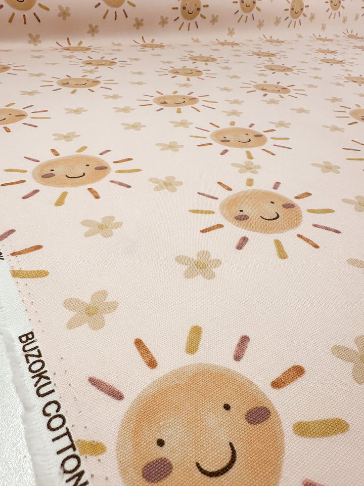 This medium-weight cotton duck fabric, "Sunny" from Super Cheap Fabrics, showcases smiling suns with pastel rays and scattered flowers on a light pink background. The cheerful design is perfect for home décor. Available in 112cm width.