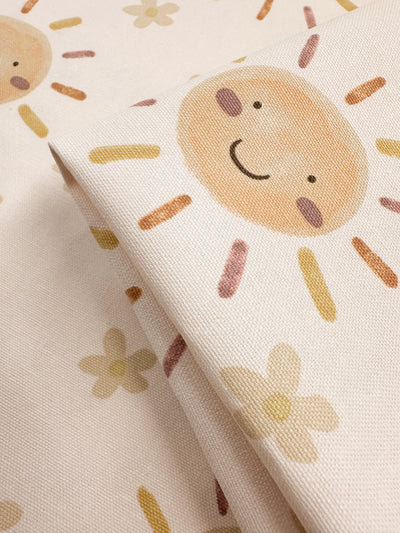 The Super Cheap Fabrics' Cotton Duck - Sunny - 112cm is ideal for home décor, featuring a cheerful pattern of smiling suns with rosy cheeks and vibrant rays, alongside simple flowers in warm colors. The medium-weight fabric showcases the charming design through its beautiful repetition.