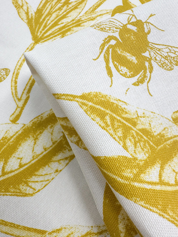 Close-up of Super Cheap Fabrics' Cotton Duck - Bee Mustard (150cm) showing its intricate print of bees and leaves on spicy mustard hue cotton fabric. The folded material highlights texture, ideal for adding a touch of nature to home décor.