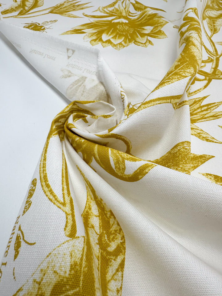 A close-up of Super Cheap Fabrics' Cotton Duck in Bee Mustard features a white cotton with a spicy mustard floral pattern, draped elegantly. The intricate botanical elements highlight the fabric's texture and design, ideal for home décor. Width: 150cm.