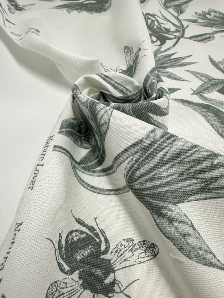This medium-weight cotton duck fabric, "Bee Seagrass" by Super Cheap Fabrics, features green botanical and insect illustrations with detailed leaves and a bee on a slightly wrinkled texture. Its elegant "Nature Lover" print adds charm, making it ideal for home décor. Width: 150cm.