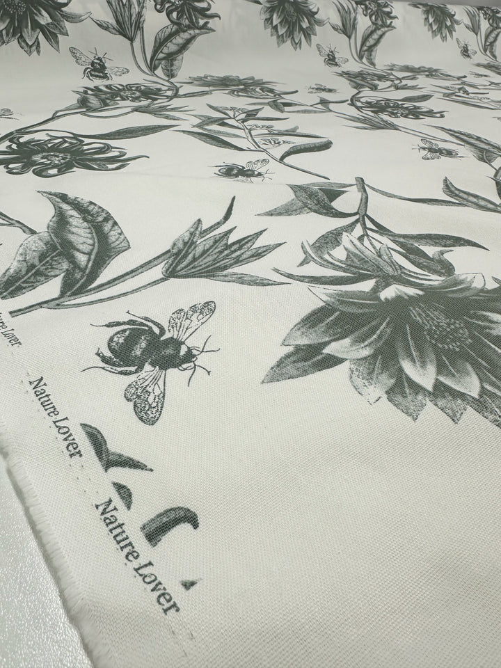 Super Cheap Fabrics' Cotton Duck - Bee Seagrass is a medium weight cotton featuring a black and white nature-themed design with flowers, bees, and leaves. The words "Nature Lover" elegantly adorn its edge, making it perfect for home décor projects. Width: 150cm.