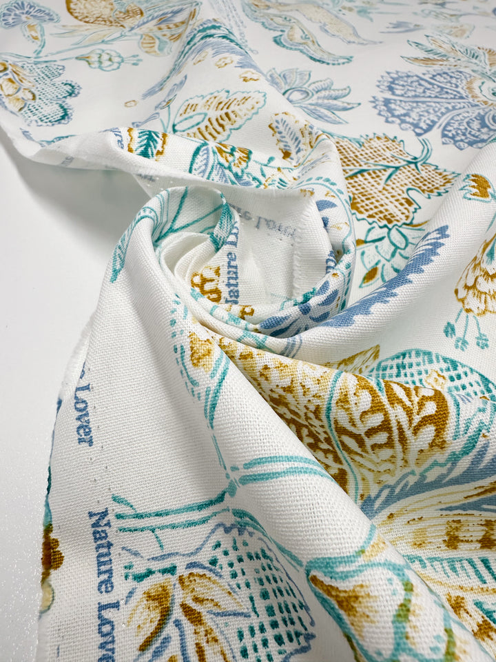 A close-up of the medium weight Cotton Duck - Fine Lines fabric by Super Cheap Fabrics shows a white backdrop with a teal, mustard, and blue floral pattern. Perfect for home décor, it artistically features "Nature Lover" in blue within gentle folds.
