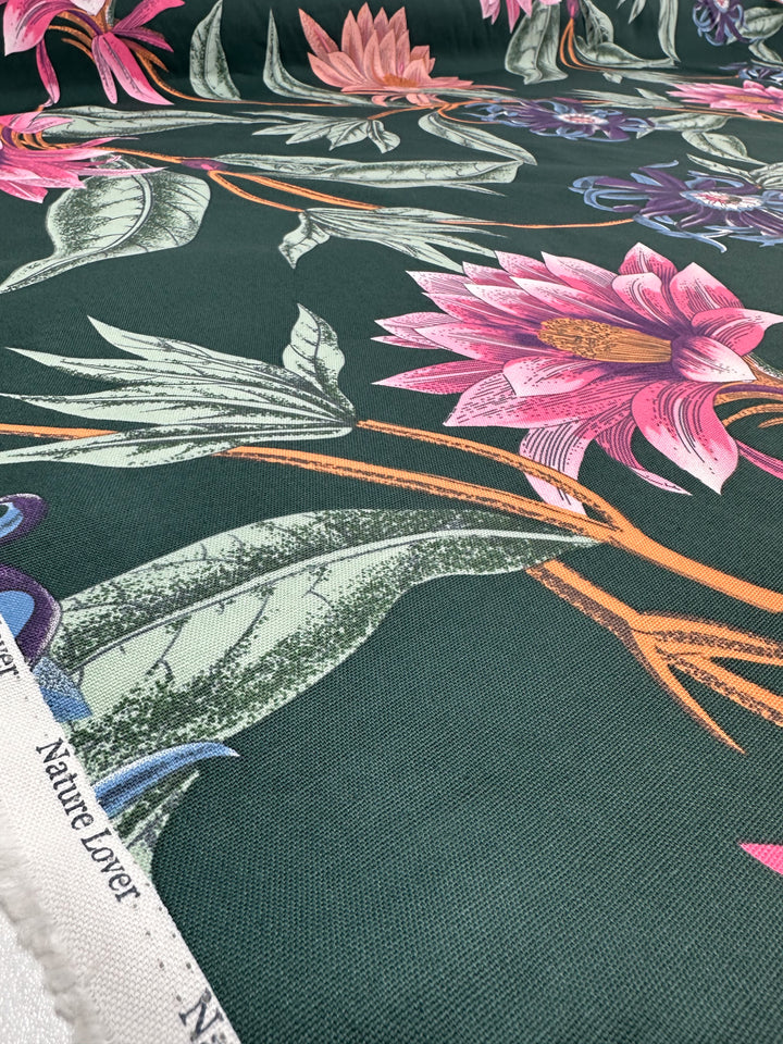 Close-up of Super Cheap Fabrics' Cotton Duck - Tropical Punch, a medium weight fabric with a dark green background adorned with large pink and purple flowers and green leaves. "Nature Lover" is elegantly displayed on the fabric's edge, perfect for home décor. Width: 147cm.