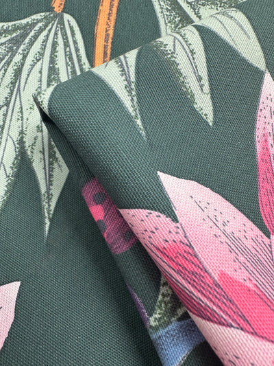 Close-up of folded Super Cheap Fabrics' Cotton Duck - Tropical Punch, perfect for home décor. This medium weight fabric showcases large green leaves and pink flowers on a dark green background, revealing intricate leaf and petal detailing with its visible texture. Width: 147cm.
