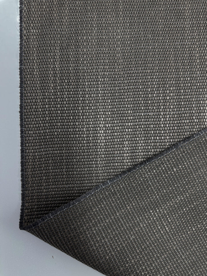 A close-up of Grid Upholstery - Yari 117 – 147cm by Super Cheap Fabrics in textured dark gray, similar to lava smoke, is shown on a light gray background. The weave and flexibility of this heavy-weight fabric are visible with one corner slightly lifted, emphasizing its high durability.