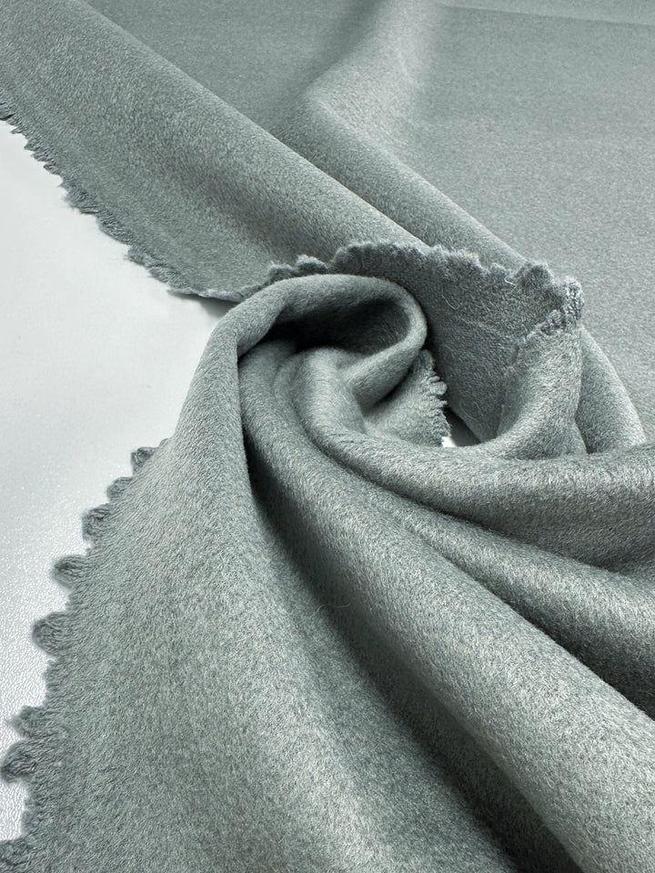 A close-up of the soft, iceberg green Deadstock Wool Cashmere from Super Cheap Fabrics, made in Italy with fringed edges, elegantly draped and twisted on a flat surface to highlight its texture and softness.