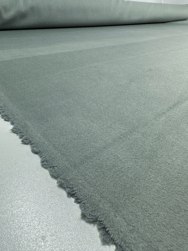 A close-up of Super Cheap Fabrics' Deadstock Wool Cashmere in Iceberg Green, 150cm wide, unfurled on a smooth surface. This Italian-made fabric features a soft texture with slightly frayed edges extending into the distance.
