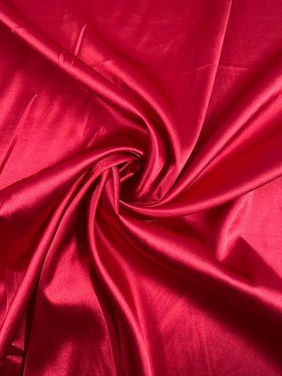 The Scarlet Smile from Super Cheap Fabrics is a vibrant red satin, expertly gathered and twisted to highlight its smooth, shimmering texture. The deluxe material's central swirl elegantly reflects light on its silky surface.