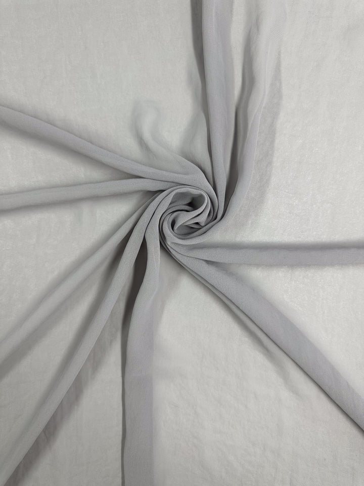 The Hi-Multi Chiffon - Harbor Mist by Super Cheap Fabrics features sheer gray fabric twisted into a spiral, forming a star-like pattern with soft folds extending outward on a flat surface.