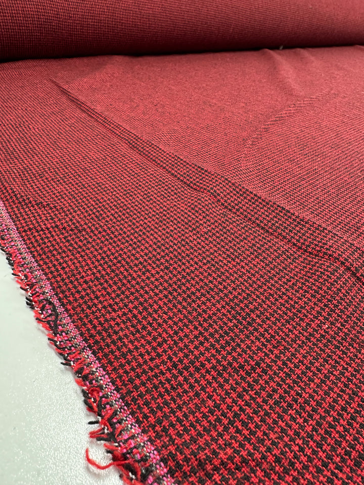 Close-up of a roll of Designer Tweed - Houndy - 155cm, a red and black houndstooth pattern by Super Cheap Fabrics. Perfect for outer coats, the medium-weight polyester fabric lays unrolled on a flat surface with prominently visible texture and frayed edges.