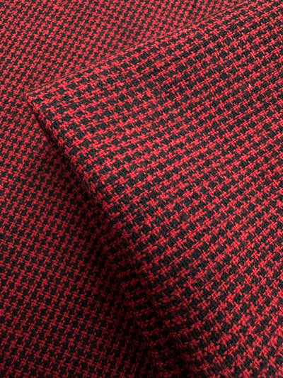 A close-up of the Super Cheap Fabrics' Designer Tweed - Houndy - 155cm showcases a red and black houndstooth polyester blend. The medium-weight fabric, perfect for outer coats, features small, broken checks in alternating colors and appears folded to reveal its texture and intricate design.