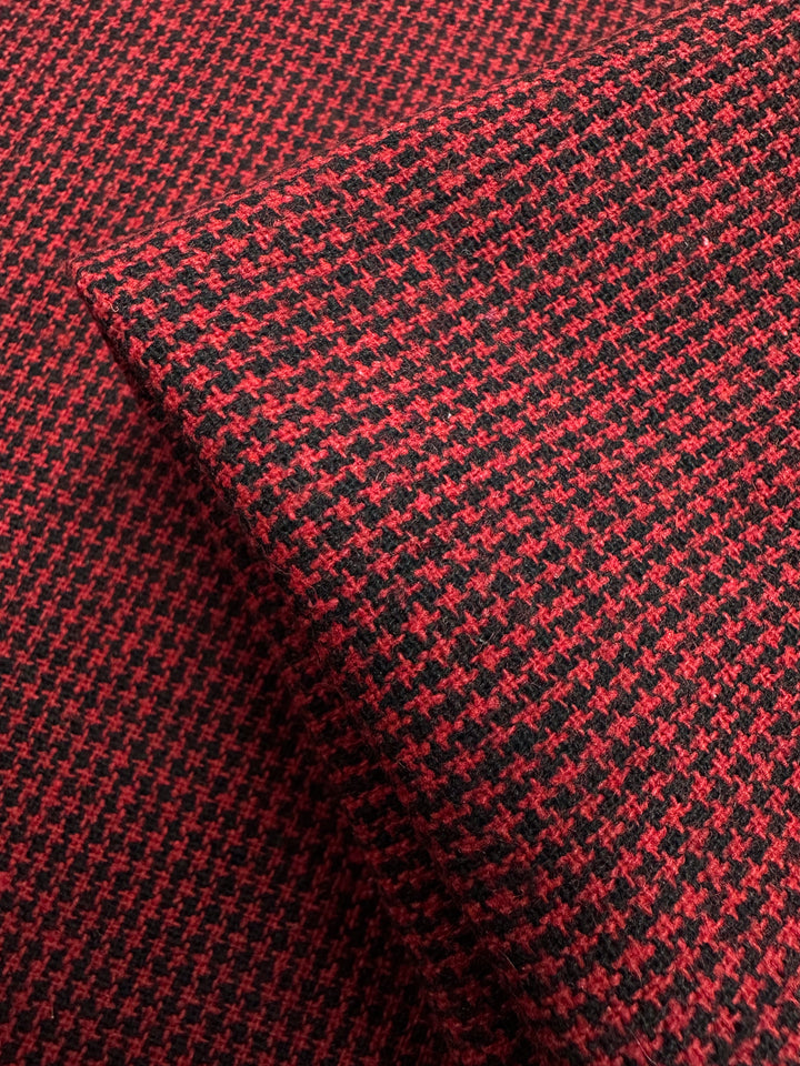 A close-up of the Super Cheap Fabrics' Designer Tweed - Houndy - 155cm showcases a red and black houndstooth polyester blend. The medium-weight fabric, perfect for outer coats, features small, broken checks in alternating colors and appears folded to reveal its texture and intricate design.
