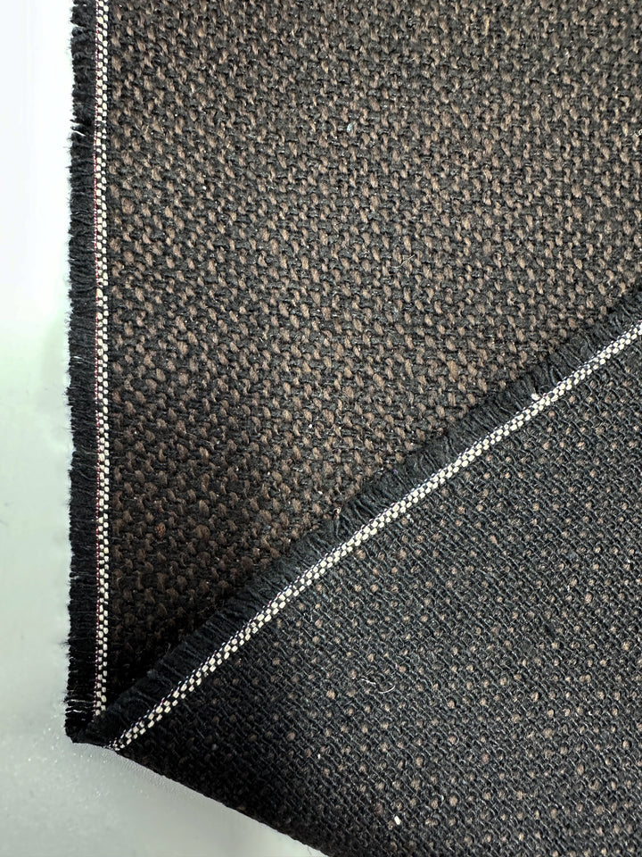 Close-up of a dark, heavyweight Designer Tweed - Choco by Super Cheap Fabrics, showcasing a visible selvage with white and red stripes. The 150cm folded polyester fabric reveals both sides, highlighting its woven pattern and rich texture—perfect for crafting outer coats.