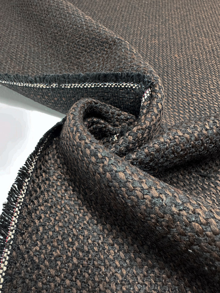 A close-up of the Designer Tweed - Choco by Super Cheap Fabrics reveals a textured, woven polyester fabric in dark brown with hints of lighter brown. A slightly folded edge shows a stitched border featuring small white, pink, and green dots. This heavy-weight fabric is soft and thick, ideal for outer coats.