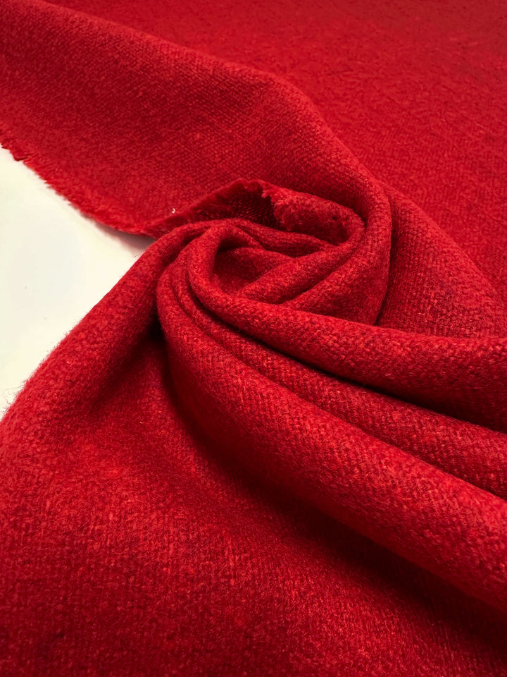 A close-up of Super Cheap Fabrics' Brushed Tweed in "Flame" reveals soft polyester folded into a spiral on a light surface. The fabric's thick, woolly texture and vivid scarlet hue catch the light, enhancing its rich color and softness. Width: 150cm.