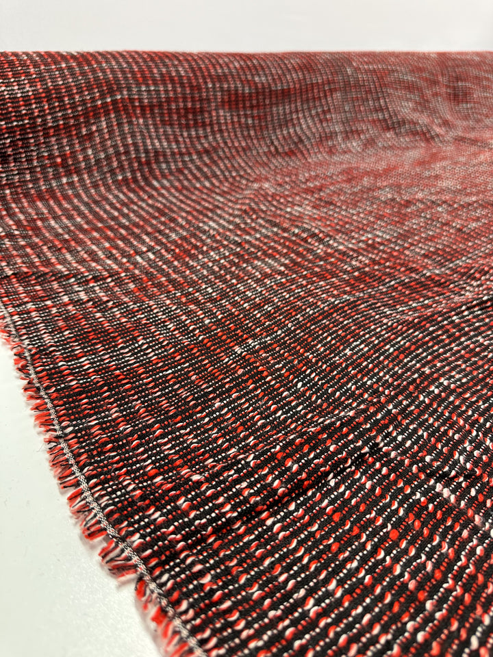 A close-up of Designer Tweed - Ripple Red - 157cm by Super Cheap Fabrics shows its textured red and black medium weight fabric with fringe edges, perfect for jackets. Spread out on a white surface, the material displays its detailed pattern and gradient coloration.