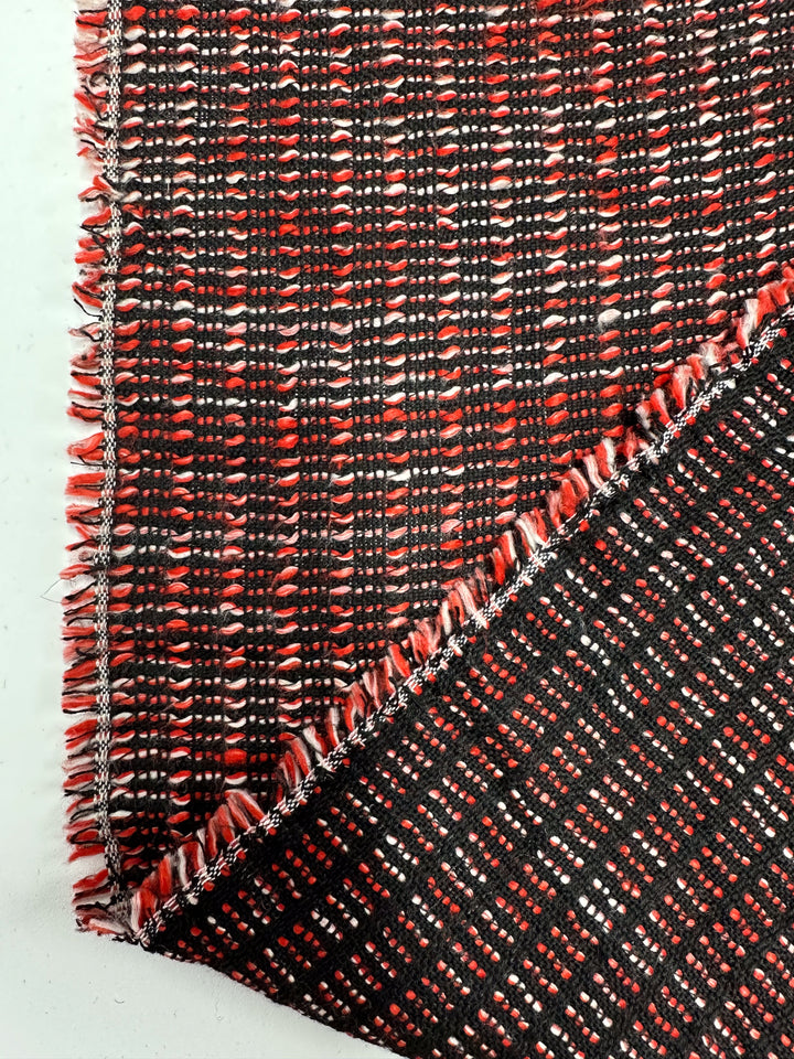 A close-up of Super Cheap Fabrics' Designer Tweed - Ripple Red - 157cm reveals a textured red-and-black pattern, perfect for jackets or coats. The medium-weight material is folded at the corner, highlighting its loose fringe and grid-like design with alternating rows.