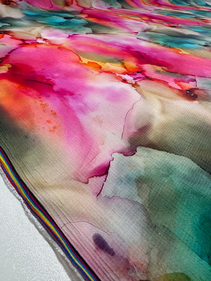 Close-up of Designer Cotton - Monique by Super Cheap Fabrics, featuring vibrant, abstract watercolor patterns in pink, red, teal, and orange hues with a fluid marbled texture. A striped edge adorns the bottom left corner.