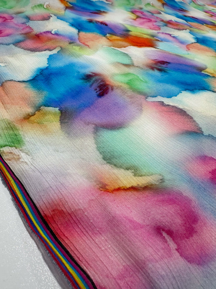 A close-up of Super Cheap Fabrics' Designer Cotton - Airbrush (150cm) displays a seamless blend of blue, pink, purple, and green hues on textured fabric. The edge is adorned with a thin rainbow stripe, ideal for luxury sewing projects.