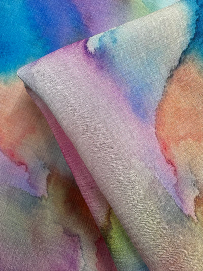 Close-up of Super Cheap Fabrics' Designer Cotton - Airbrush (150cm) featuring a vibrant watercolor pattern in blue, pink, purple, and green. Softly folded to reveal its gentle texture and luxurious abstract design, ideal for sewing projects.